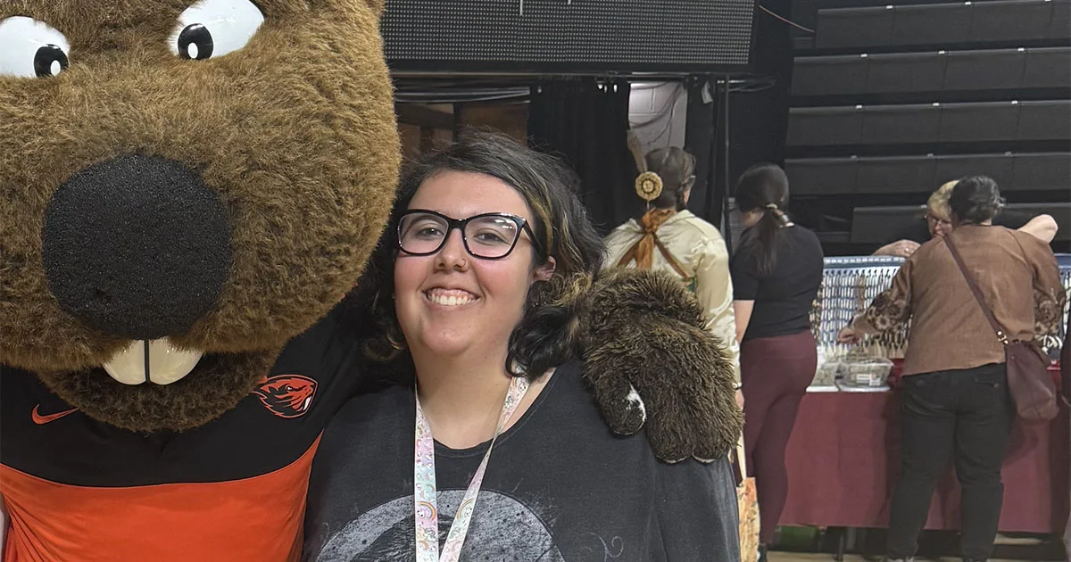 Attendee at an indoor event posing with a beaver mascot, with booths and other people in the background.