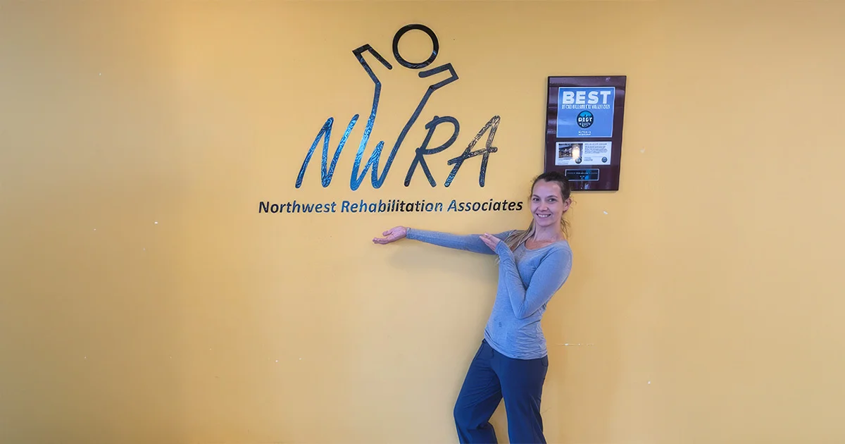 Smiling person standing beside a wall display for Northwest Rehabilitation Associates, pointing toward the organization’s logo.