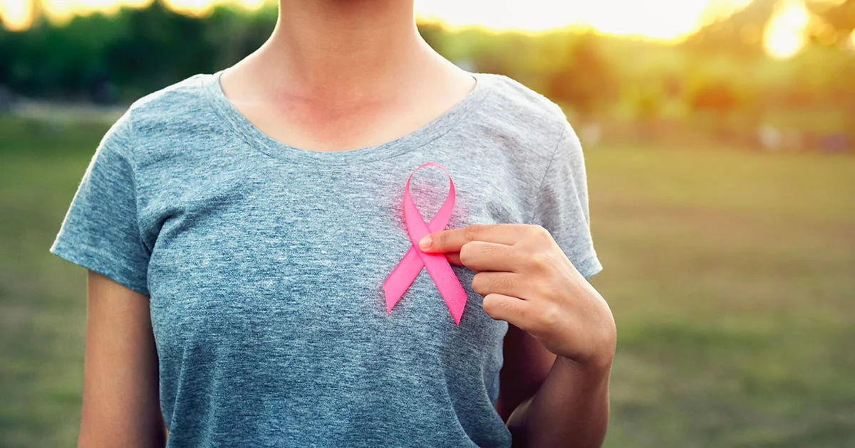 Woman wearing a gray shirt with a pink ribbon on her chest. Hand gently touches the ribbon.